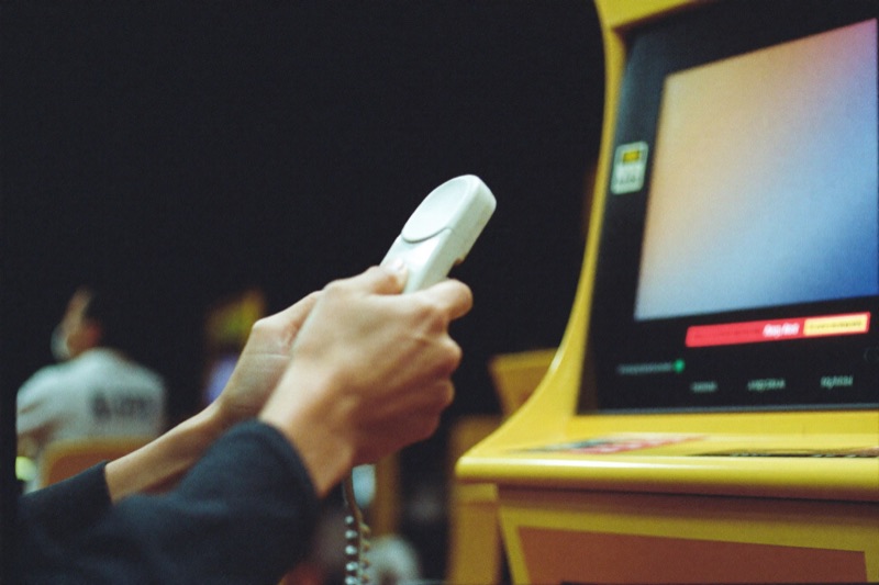 A phone held in both hands inside a slot hall with an install progress bar on the screen, lit cabinets and a seated player behind the hands