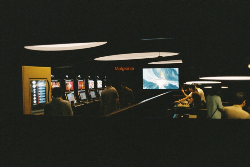 The main floor seen from a mezzanine, a bank of slot cabinets on the left and baccarat tables with dealers on the right, players walking between them under warm ceiling light