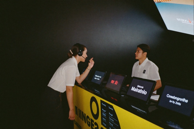 A host at a service desk inside the casino wearing a headset, leaning forward to answer a player standing at the counter