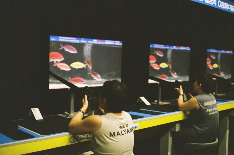 Two players side by side at a wide fish shooting cabinet, both gripping the cannon controls, bright underwater targets filling the screen in front of them