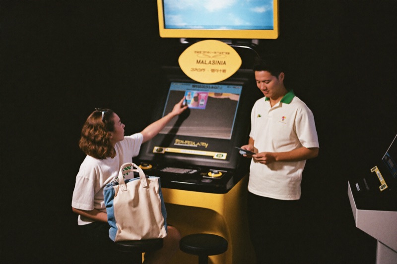 Satu pasangan muda berkongsi satu stool di slot machine, perempuan tunjuk panel bonus kecil atas screen sementara lelaki kira segenggam nota