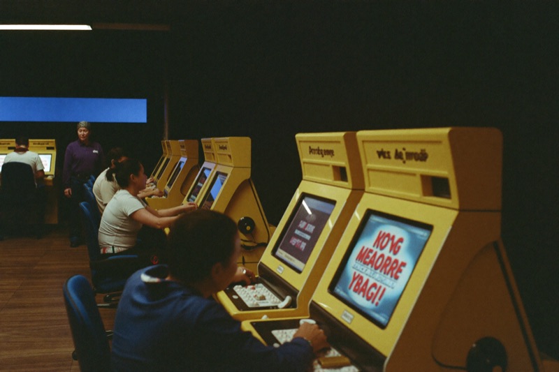 A Malaysian slot hall in the evening with a mixed group of local players at adjacent cabinets, a woman in a headscarf at the end of the row, warm lighting and a ringgit figure on one screen