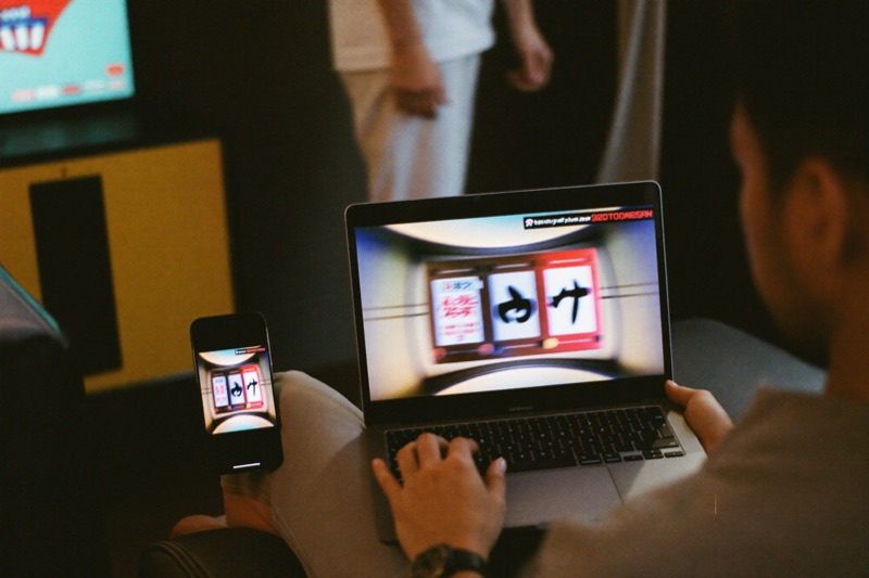 A man in his thirties on a sofa at home holding a laptop with a slot grid on the screen, a phone face up beside him showing the same reels