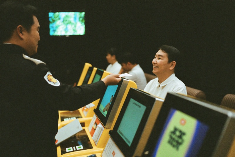 A host in a dark uniform handing a promo card to a seated slot player who looks up and smiles, two more players at machines further down the row