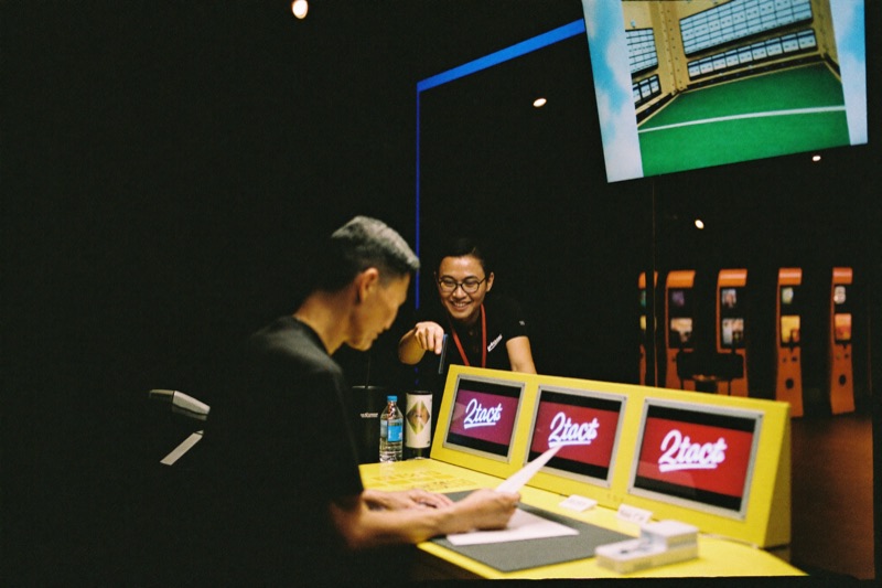 A new player at the cash desk filling in a short form while a smiling host points at the field, a slot hall visible through the glass behind them