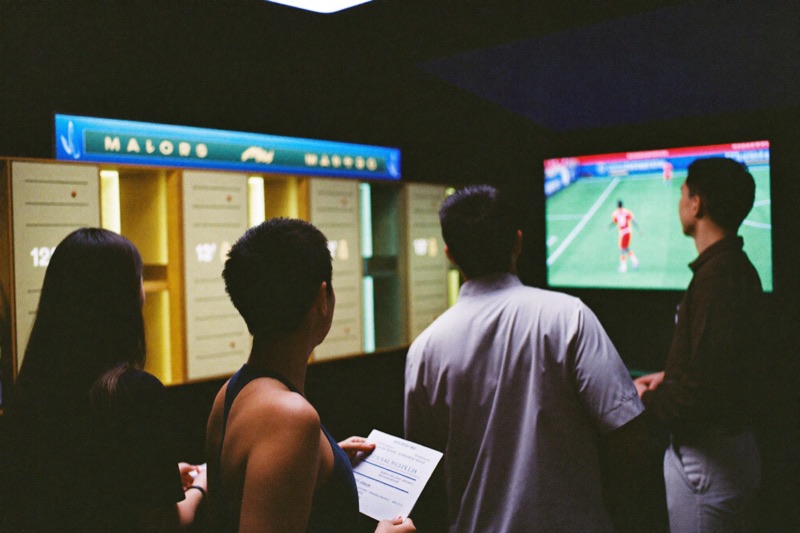 Four friends in the sports corner of a casino watching a football match on a big screen, one holding a printed slip, slot cabinets lit along the back wall