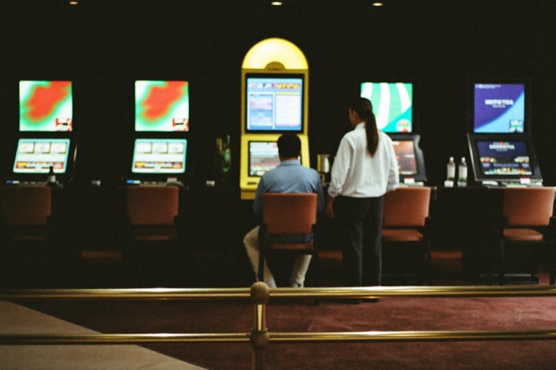 A quiet high limit slot room with four spaced cabinets, deep carpet and a brass rail, one well dressed player seated at the nearest machine with an attendant standing by