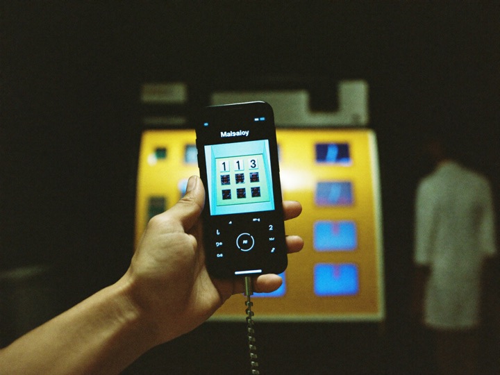 A hand holding a phone showing a slot grid, a lit slot cabinet out of focus behind it