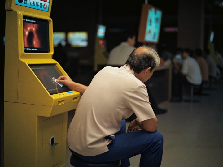 A regular player settling onto his usual stool at a slot cabinet and sliding a card into the reader, the hall busy behind him