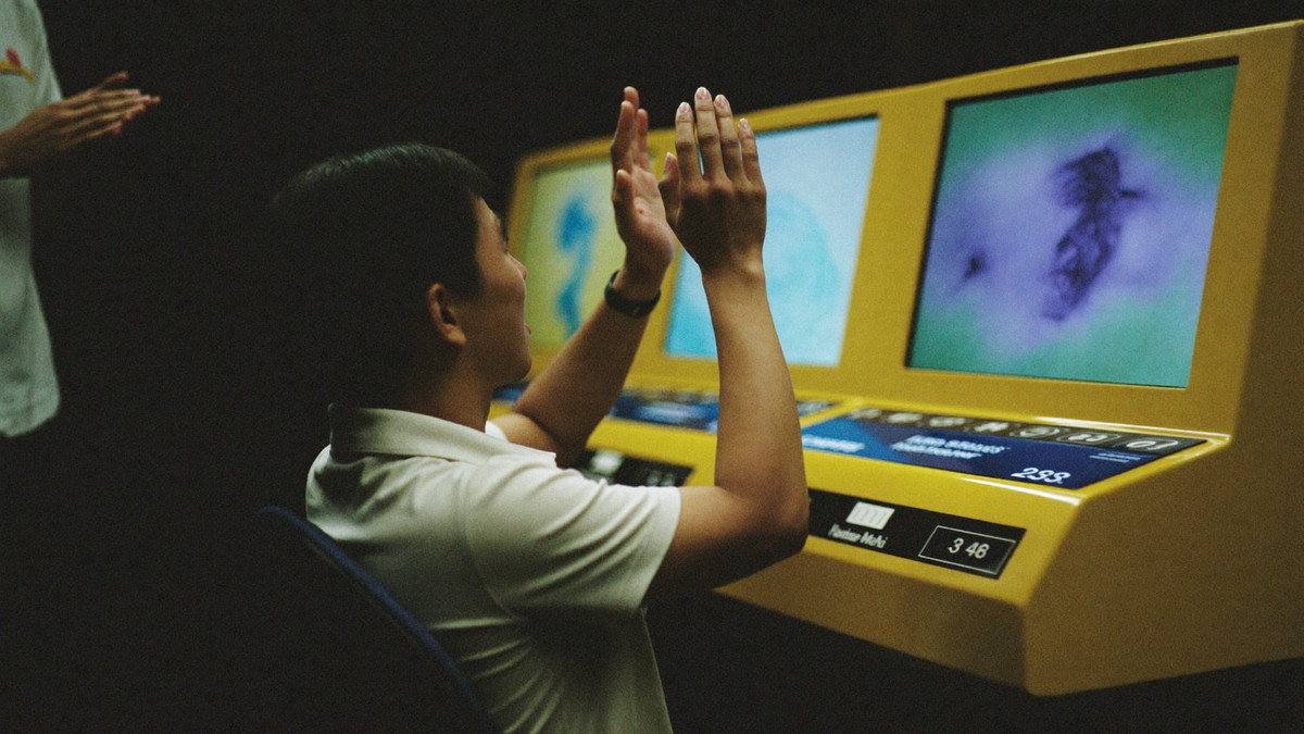 A seated slot player raising both hands as the screen in front of him lights up, a host clapping beside the cabinet